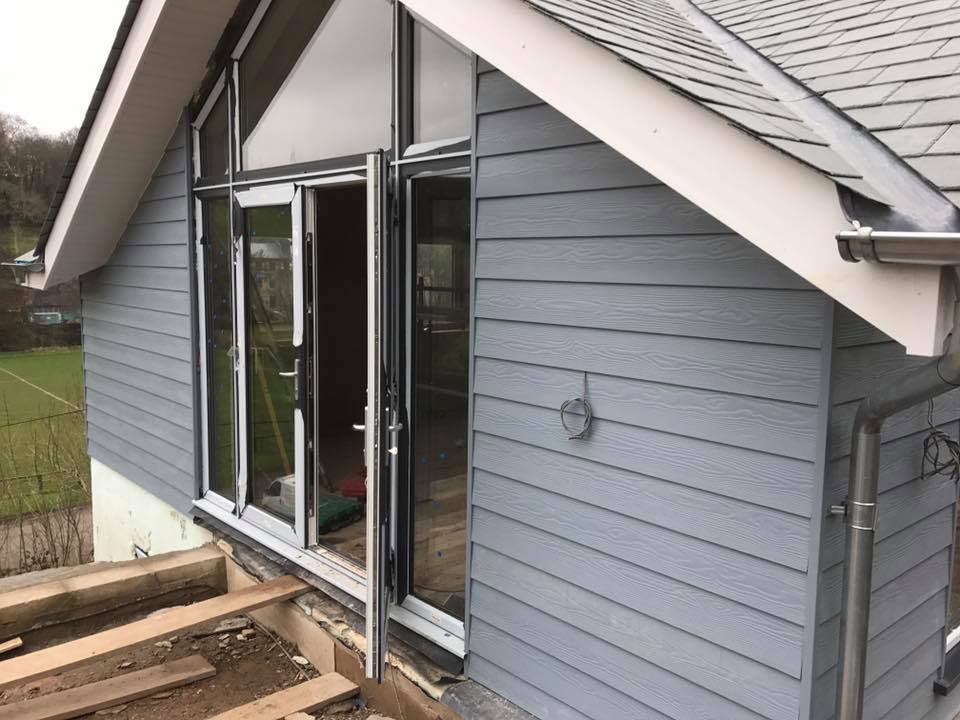 A gray house with horizontal siding, large glass doors, and exposed wooden beams on the patio area under construction. The roof has gray shingles, and the builder is focusing on kitchen installations while exterior building maintenance is ongoing.