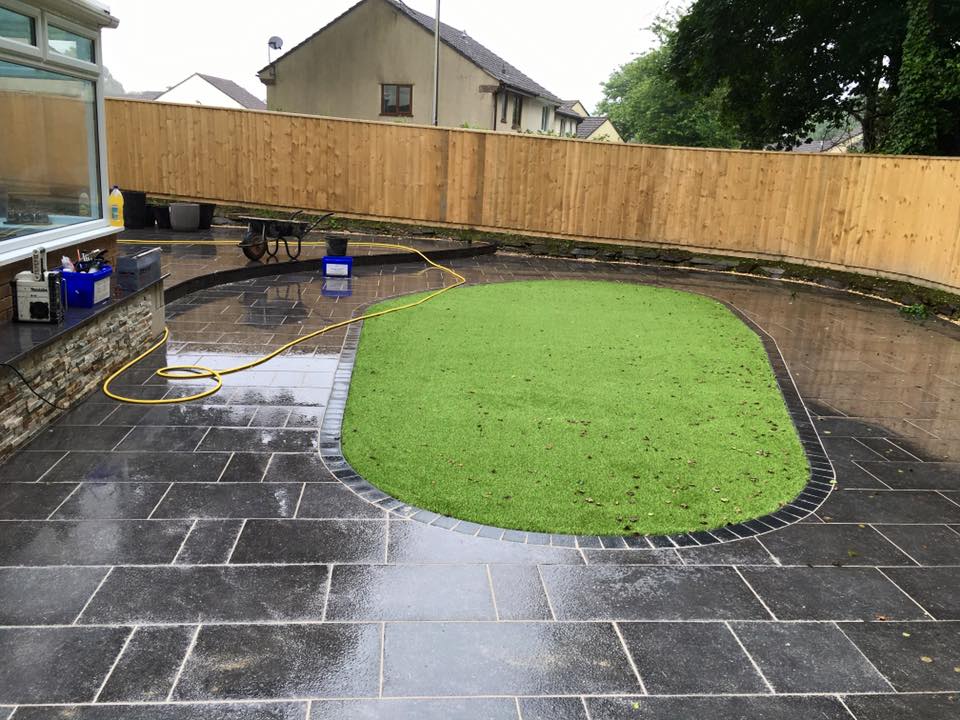 A backyard with wet black stone tiles, an oval-shaped artificial grass area in the center, a wooden fence in the background, and a yellow garden hose lying on the ground—ideal for property refurbishments or builder projects.