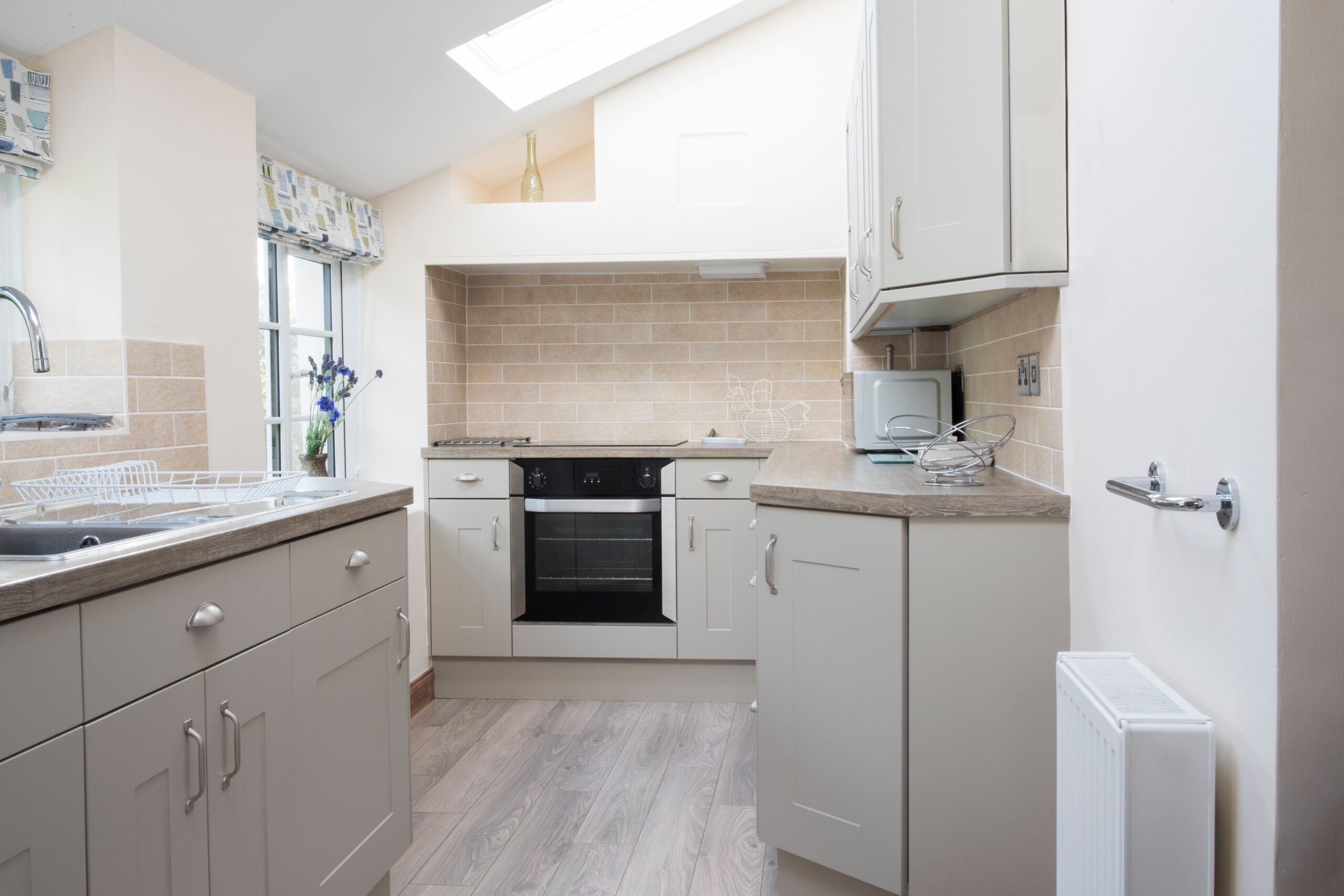 A modern, compact kitchen with light beige cabinets, built-in oven, electric stovetop, and tiled backsplash. Sunlight streams in from a skylight and windows—perfect for showcasing quality kitchen installations by an expert builder.