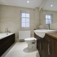 Modern bathroom with a bathtub, toilet, and large sink set in dark wood cabinets. Light-colored tiled walls and floor, a window above the toilet, and a large wall mirror showcase expert builder craftsmanship and building maintenance.