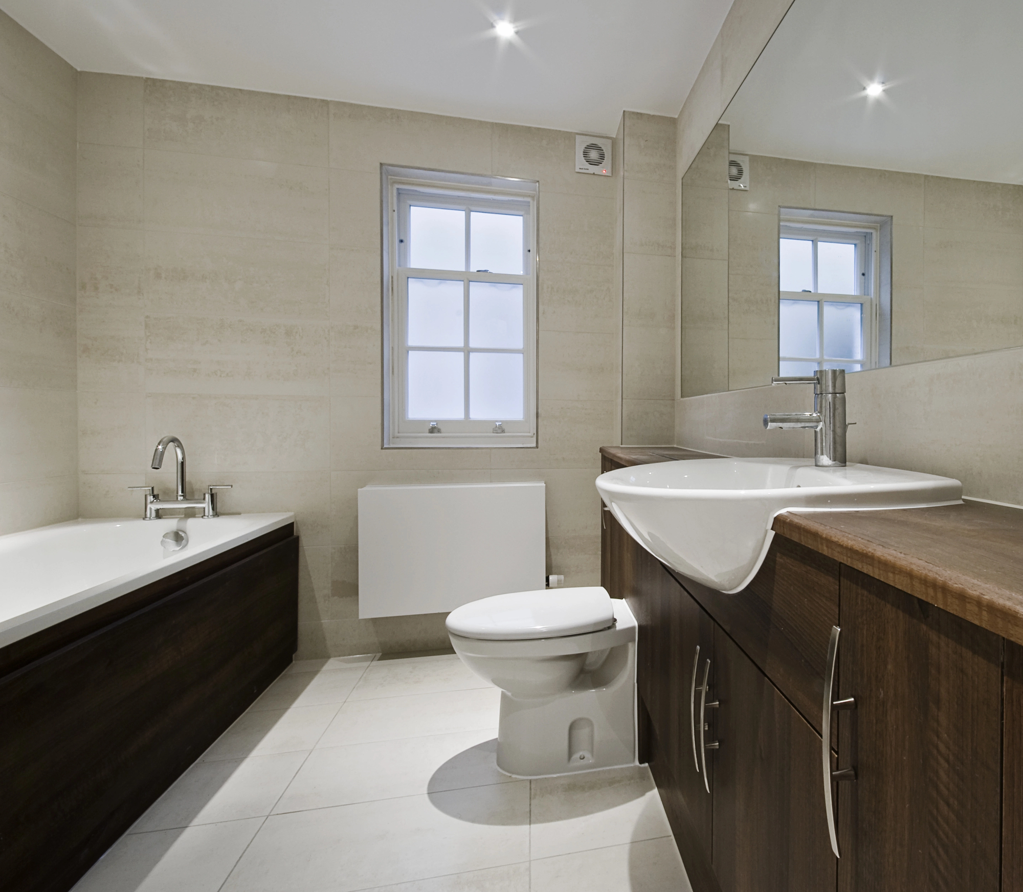 Modern bathroom with a bathtub, toilet, and large sink set in dark wood cabinets. Light-colored tiled walls and floor, a window above the toilet, and a large wall mirror showcase expert builder craftsmanship and building maintenance.