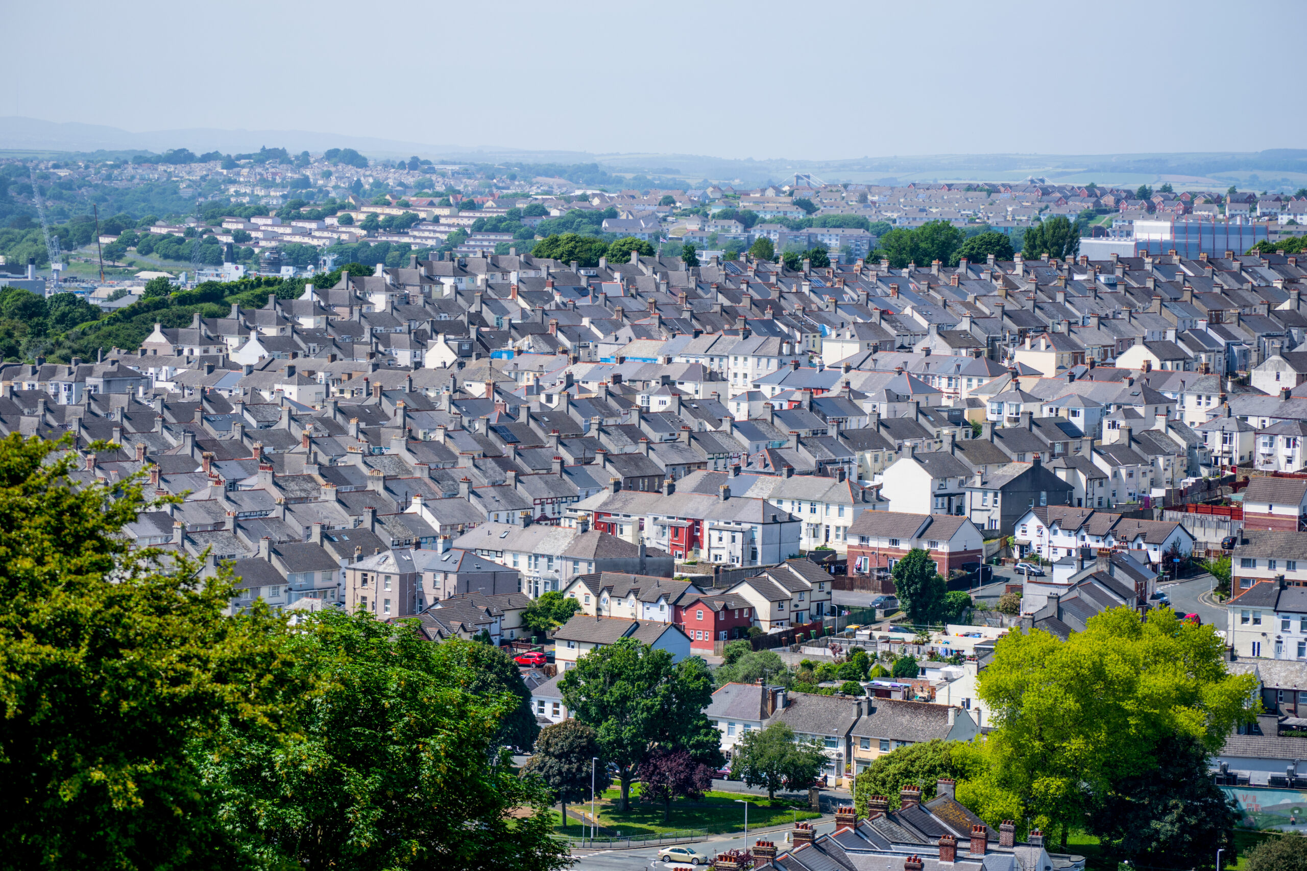 A wide view of a densely packed residential neighborhood with rows of similar houses, trees scattered throughout, and hills visible in the hazy distance—a community where property refurbishments are transforming modern living.